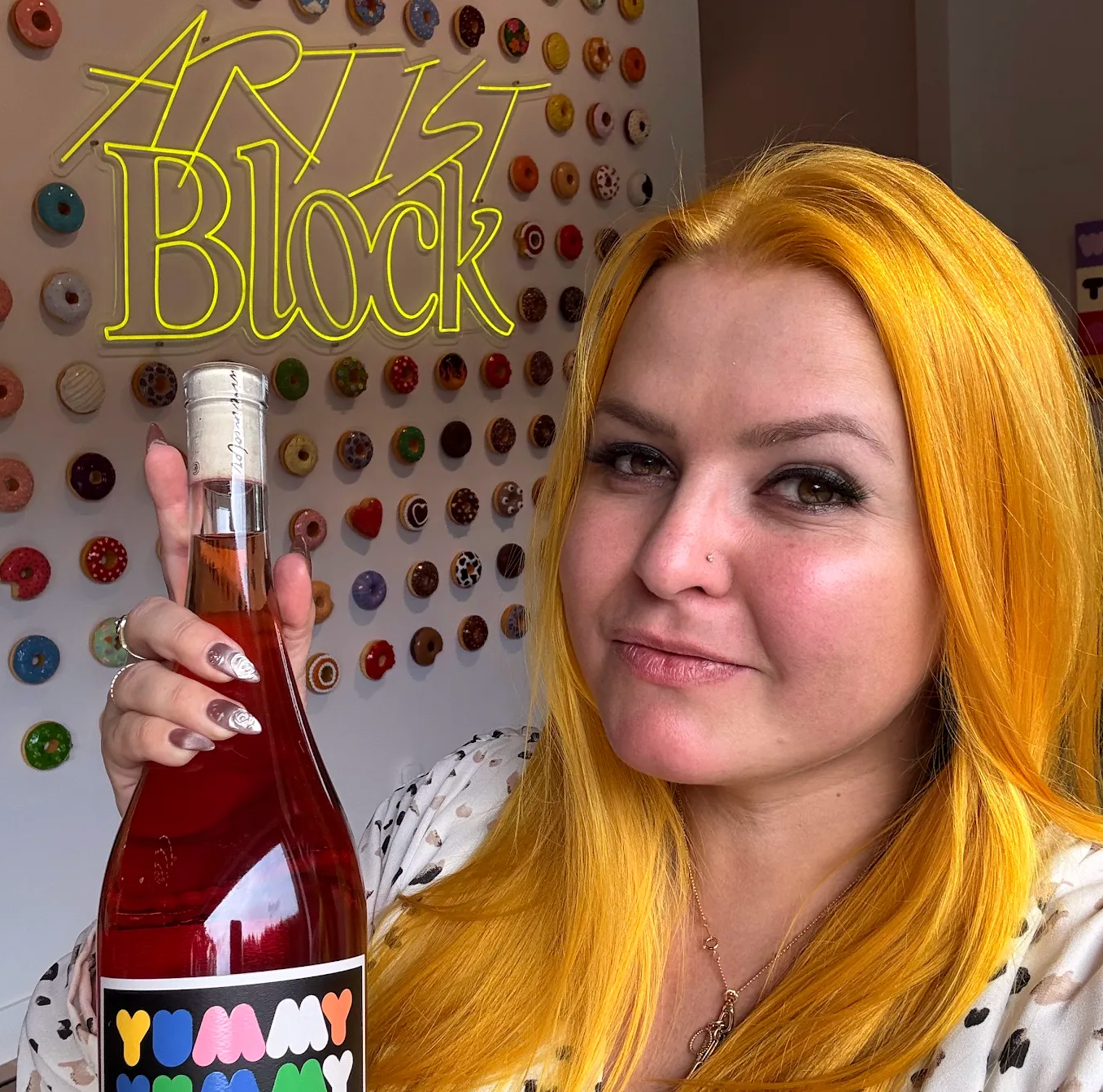 Woman with yellow hair holding a bottle labeled 'Yummy Yummy' in front of a colorful wall with 'Artist Block' text.