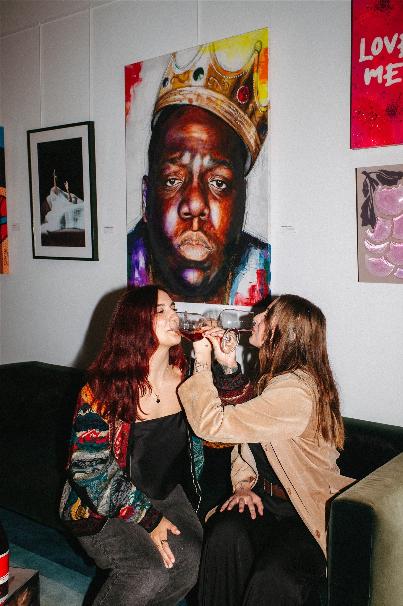 Two women sitting on a couch in front of a large painting of Biggie Smalls wearing a crown.