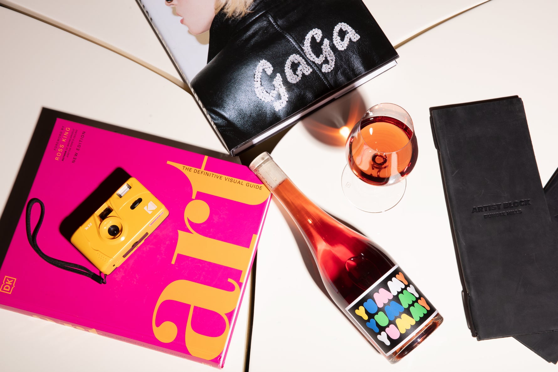 Bottle of Yummy wine with colorful label, a glass of orange wine, pink book titled 'art', and camera with 'Gaga' book.